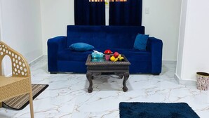 Una sala de estar con un sofá azul, una silla de madera, una mesa de centro de madera oscura con frutas y una alfombra azul.