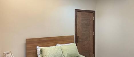 Un dormitorio con cama, cabecera de madera, mesita de noche y una lámpara, y una puerta.