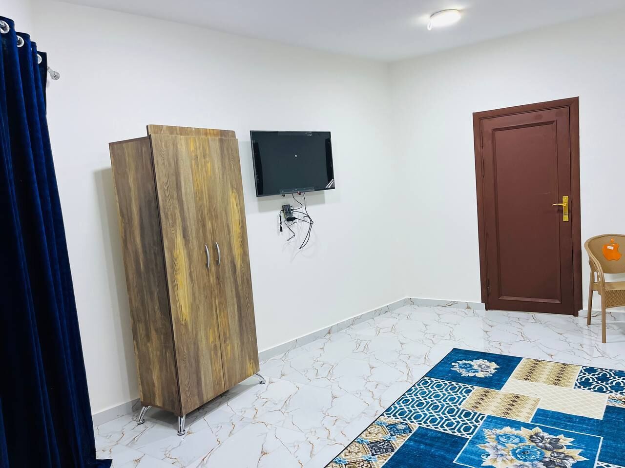 Una stanza con un armadio in legno, una TV a schermo piatto montata a parete, una porta marrone e un tappeto decorato sul pavimento.
