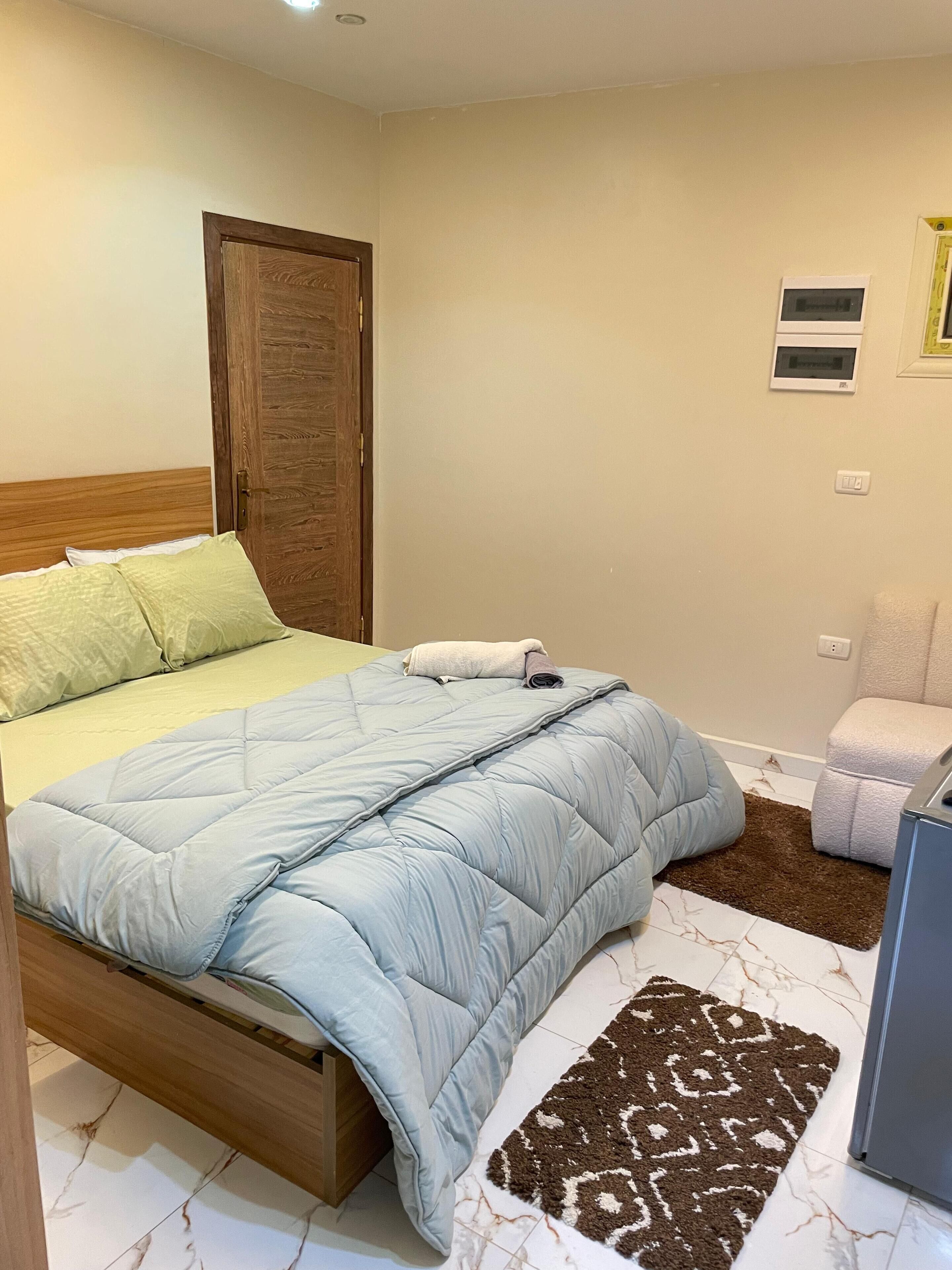 A bedroom with a bed, a wooden door, a small white chair, and a patterned rug.