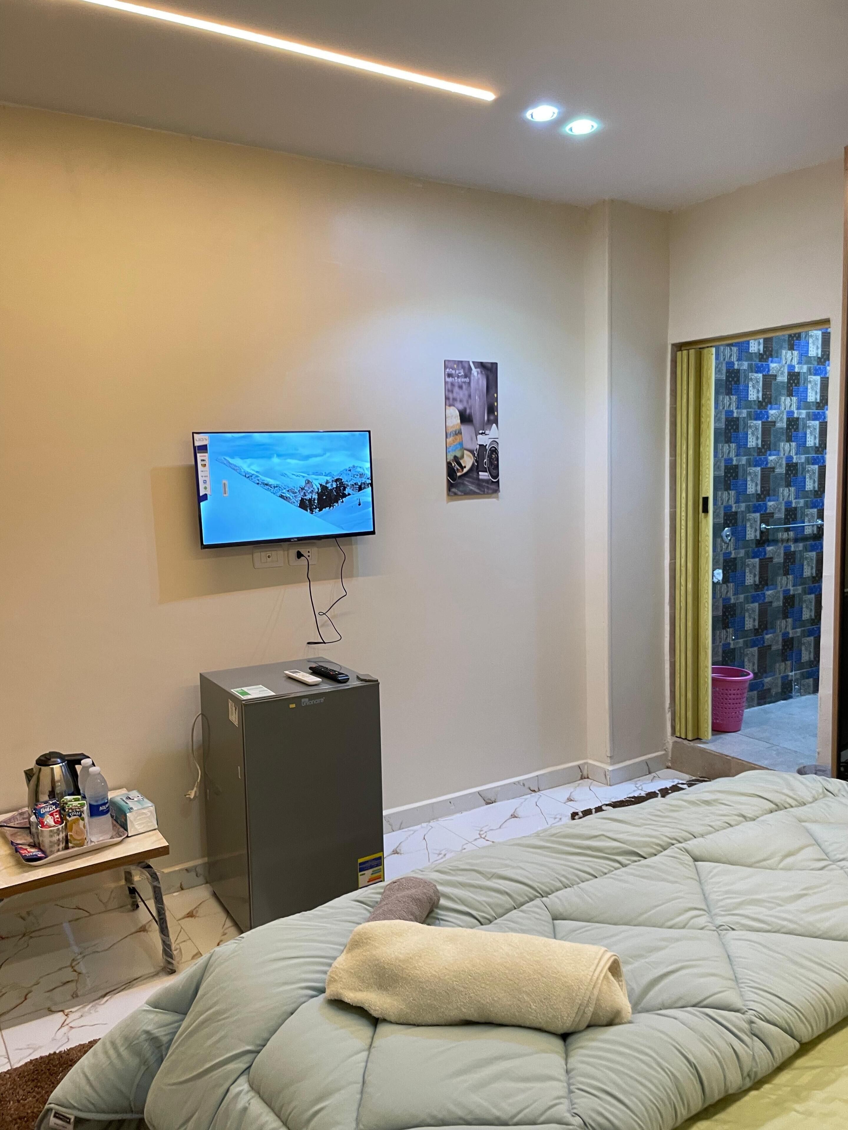 Habitación de hotel con una cama, televisor montado en la pared, un escritorio pequeño con una tetera y una botella, y una puerta con un panel de vidrio decorativo.