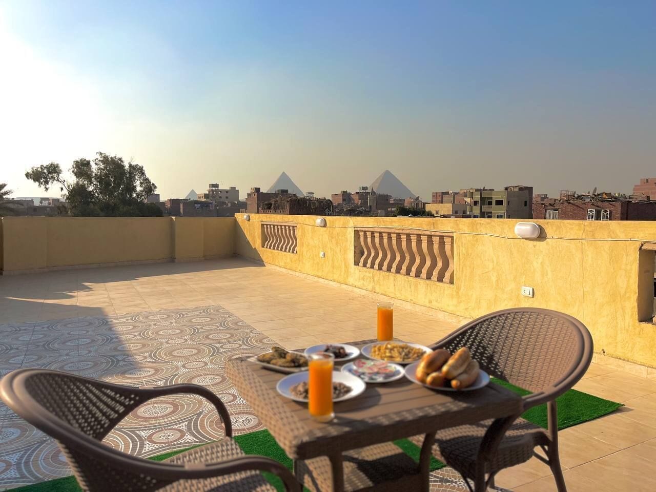 A rooftop terrace with a table set for breakfast, featuring plates of food and drinks, and a view of the pyramids in the background.