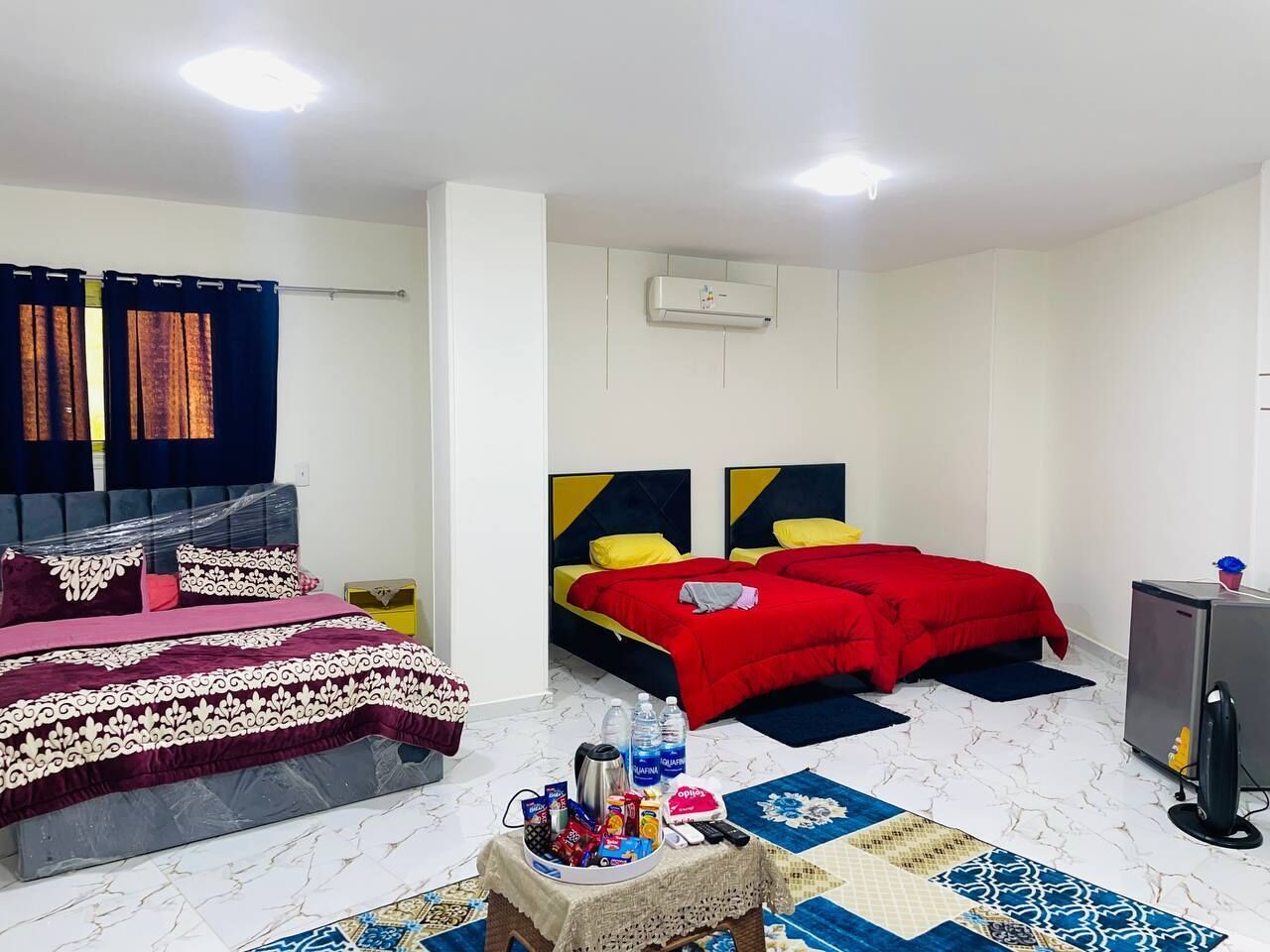 Habitación con dos camas, una con cubrecama estampado y la otra con ropa de cama roja. Hay una pequeña mesa con bocadillos y bebidas, un calentador y un dispensador de agua.