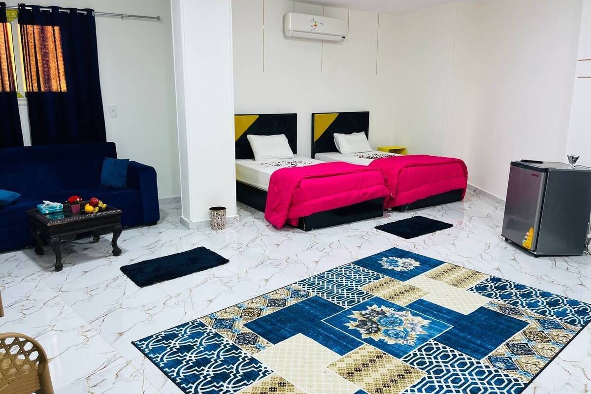 A compact room with a bed, a sofa, a small table, a refrigerator, and a patterned rug.