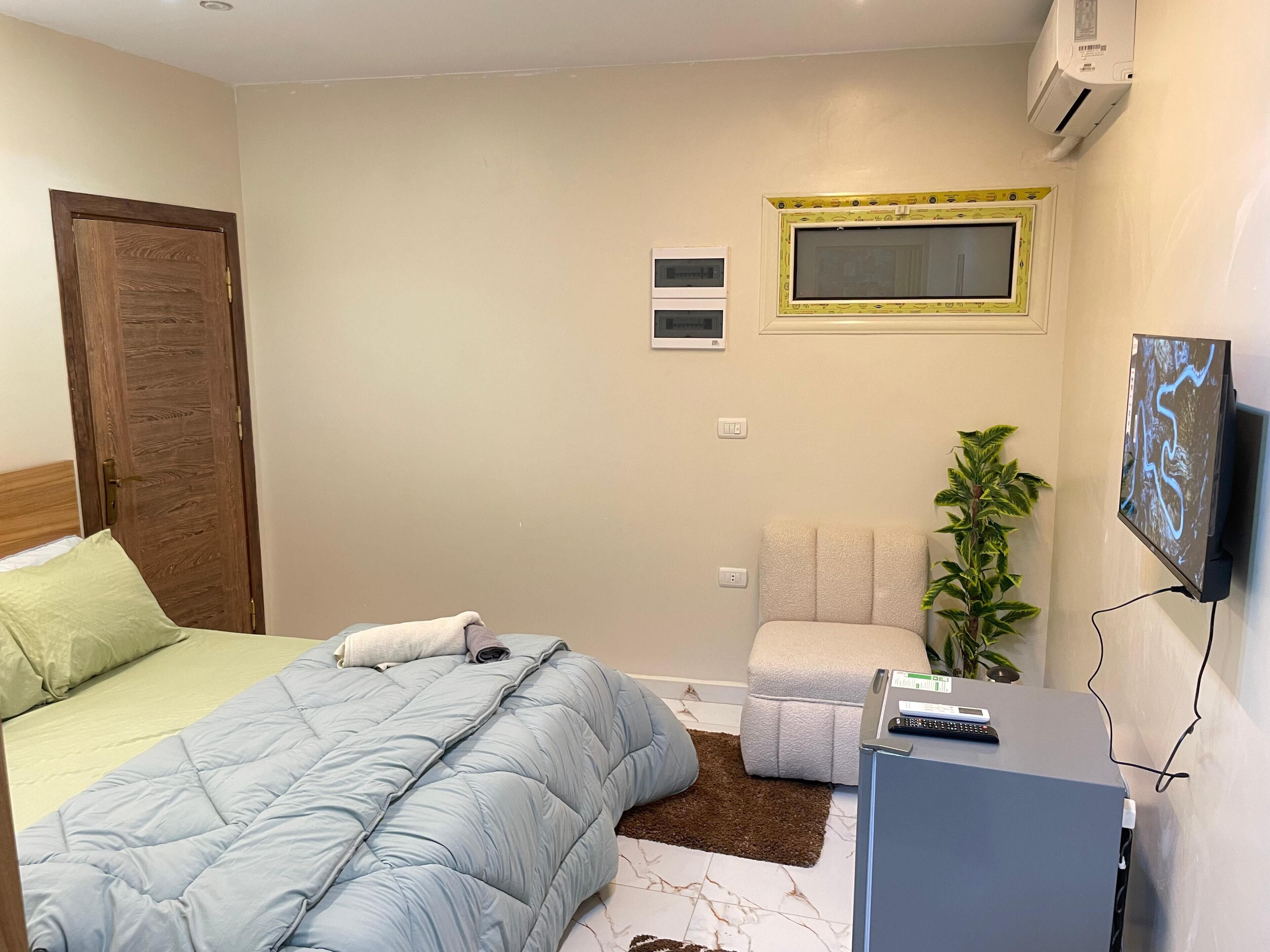 A bedroom with a bed, a desk, a chair, a potted plant, and a TV.
