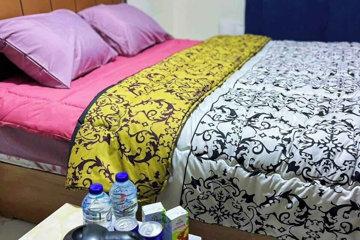 A hotel room with a bed featuring a pink and yellow blanket, a black and white patterned bedspread, and a wooden headboard. A table in the foreground has a kettle, water bottles, a juice box, and a Pepsi can.
