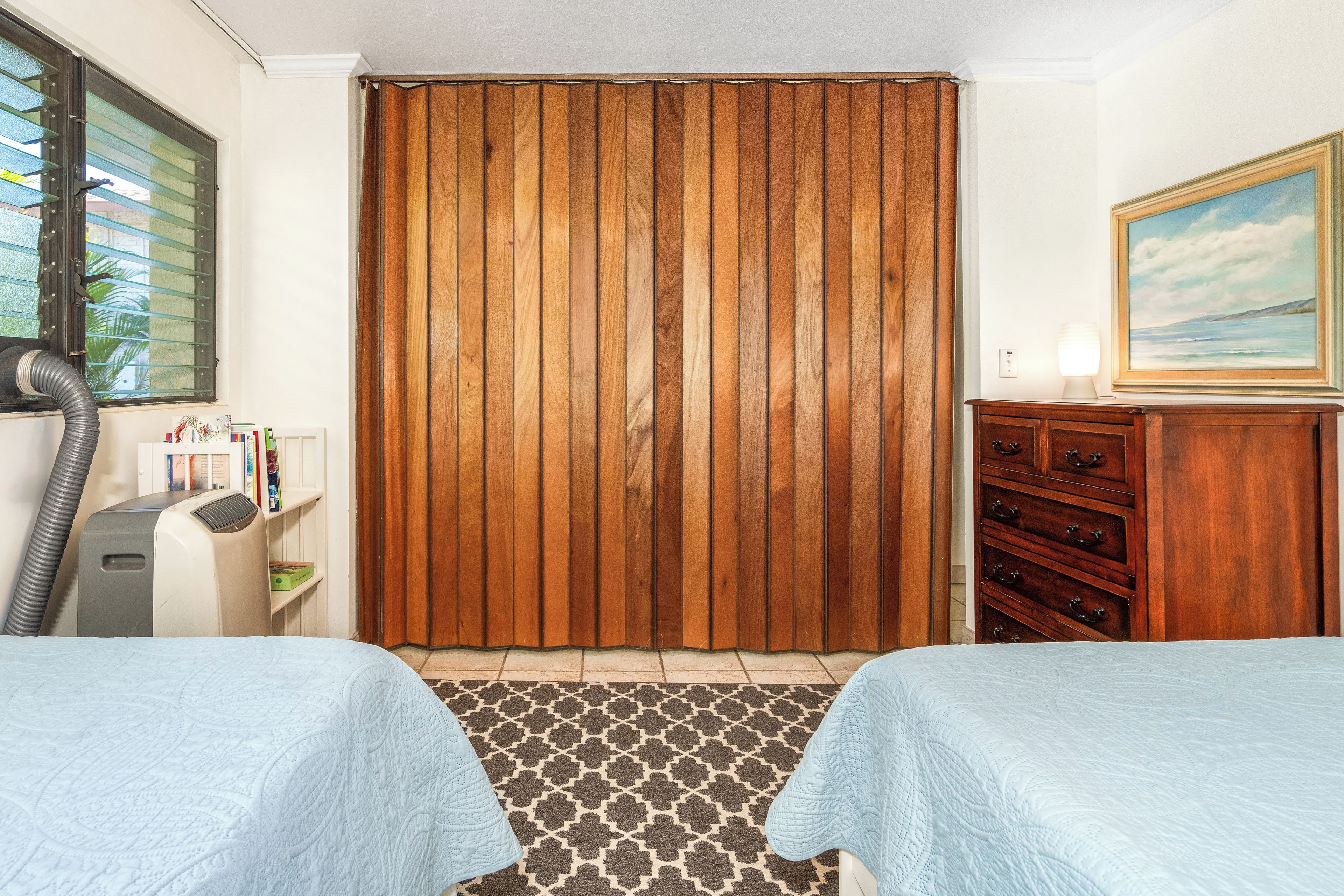 A bedroom with two beds, a wooden wardrobe, a desk with a lamp, and a painting on the wall.