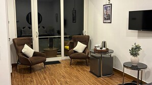 Ein modernes Hotelzimmer mit einem Bett, zwei Sesseln, einem kleinen Tisch, einem Fernseher und einer Schiebeglastür, die auf einen Balkon führt.
