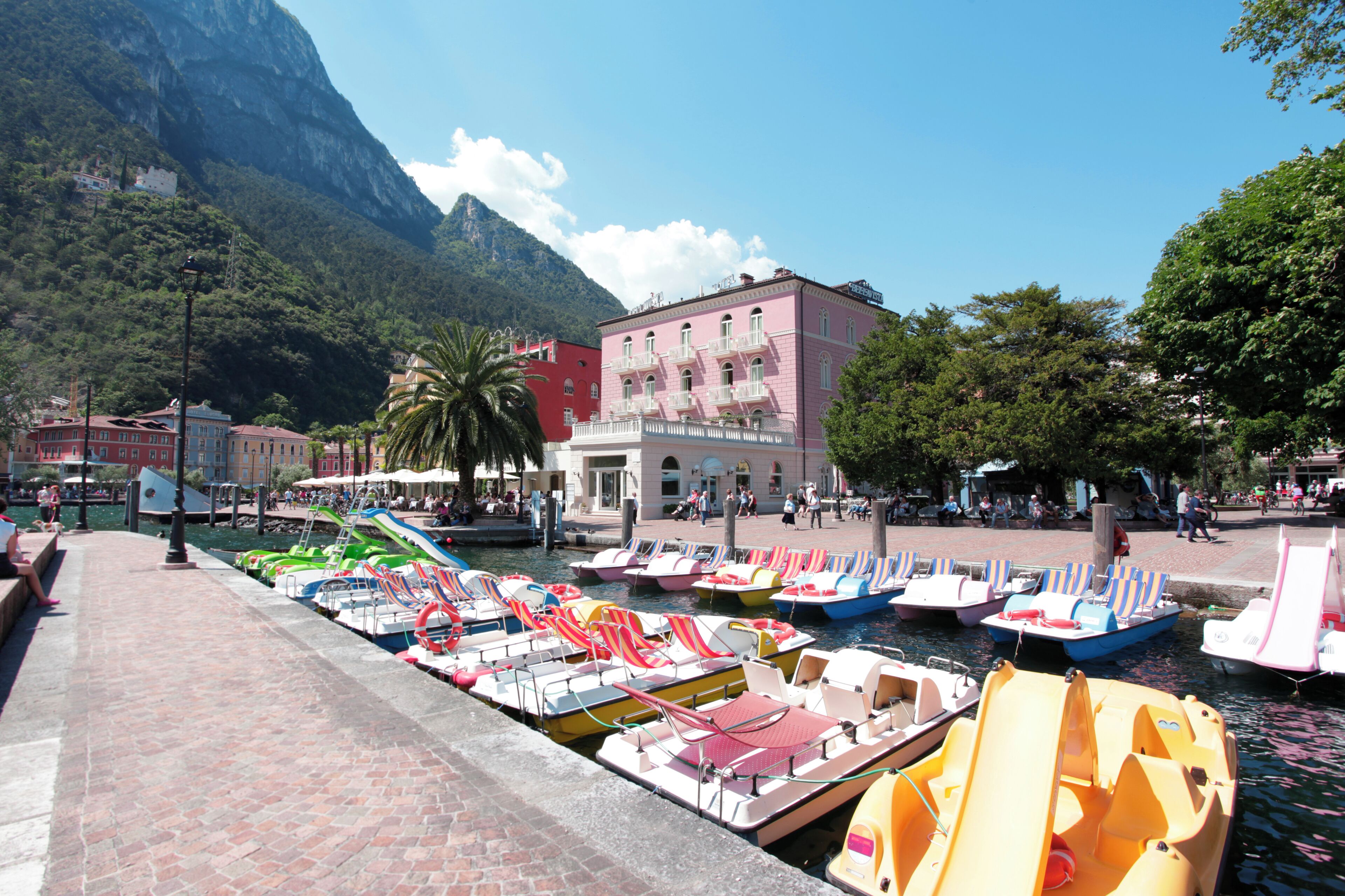 A marina with colorful boats docked, a red building with balconies, and a mountainous backdrop.