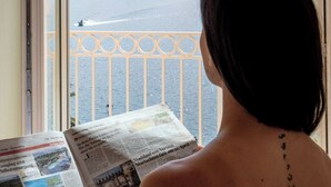 Une personne est assise près d’une fenêtre, lisant un journal, avec une vue sur une montagne et un lac.