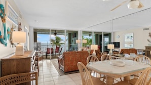 Un vaste espace de vie comprenant une table à manger, des chaises et un canapé. Un balcon offre une vue sur les palmiers et l’océan.