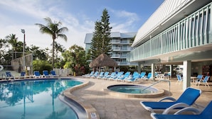 Un espace piscine avec des chaises longues et un abri de fortune à toit de chaume, entouré de palmiers et d’un immeuble à plusieurs étages doté de balcons.