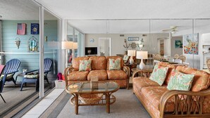 Un salon spacieux avec un grand canapé, des fauteuils en osier et une table basse en verre. On y trouve également un coin repas avec une table et des chaises, ainsi qu’une cuisine à l’arrière-plan.