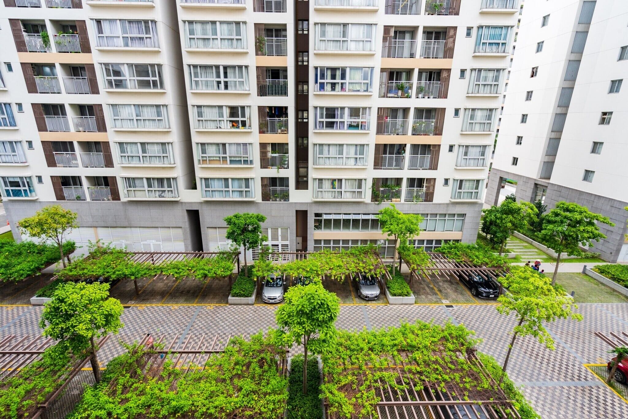 A multi-story residential building with balconies, a parking area with cars, and green landscaping.