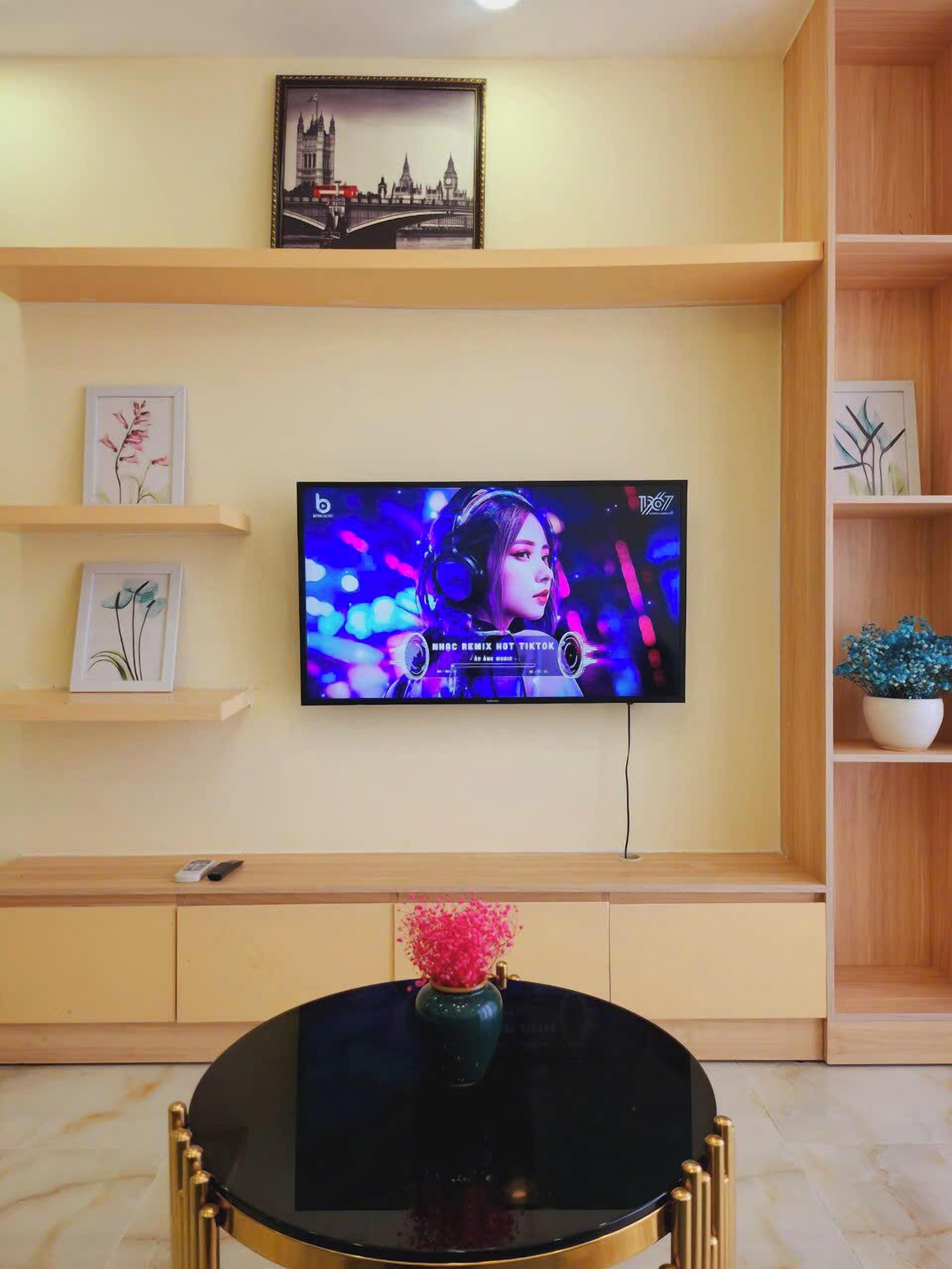 Un salon doté d’un téléviseur à écran plat fixé au mur, d’une table basse ronde sur laquelle se trouve un vase, et d’étagères en bois ornées de photos encadrées et d’une plante.