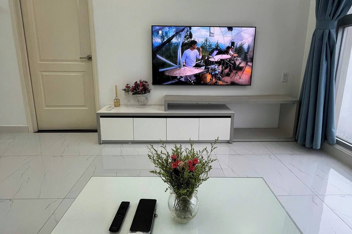 Un salon moderne avec un téléviseur à écran plat fixé au mur, une table basse blanche ornée d’un vase de fleurs et une paire de télécommandes.