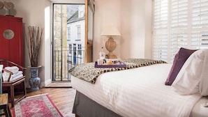 A bedroom with a bed, a nightstand, a lamp, a window with blinds, and a view of the street outside. - Upper Deck, The Upper Deck - Unique Harbourside 2Bed apartment with FREE Parking (Mevagissey)