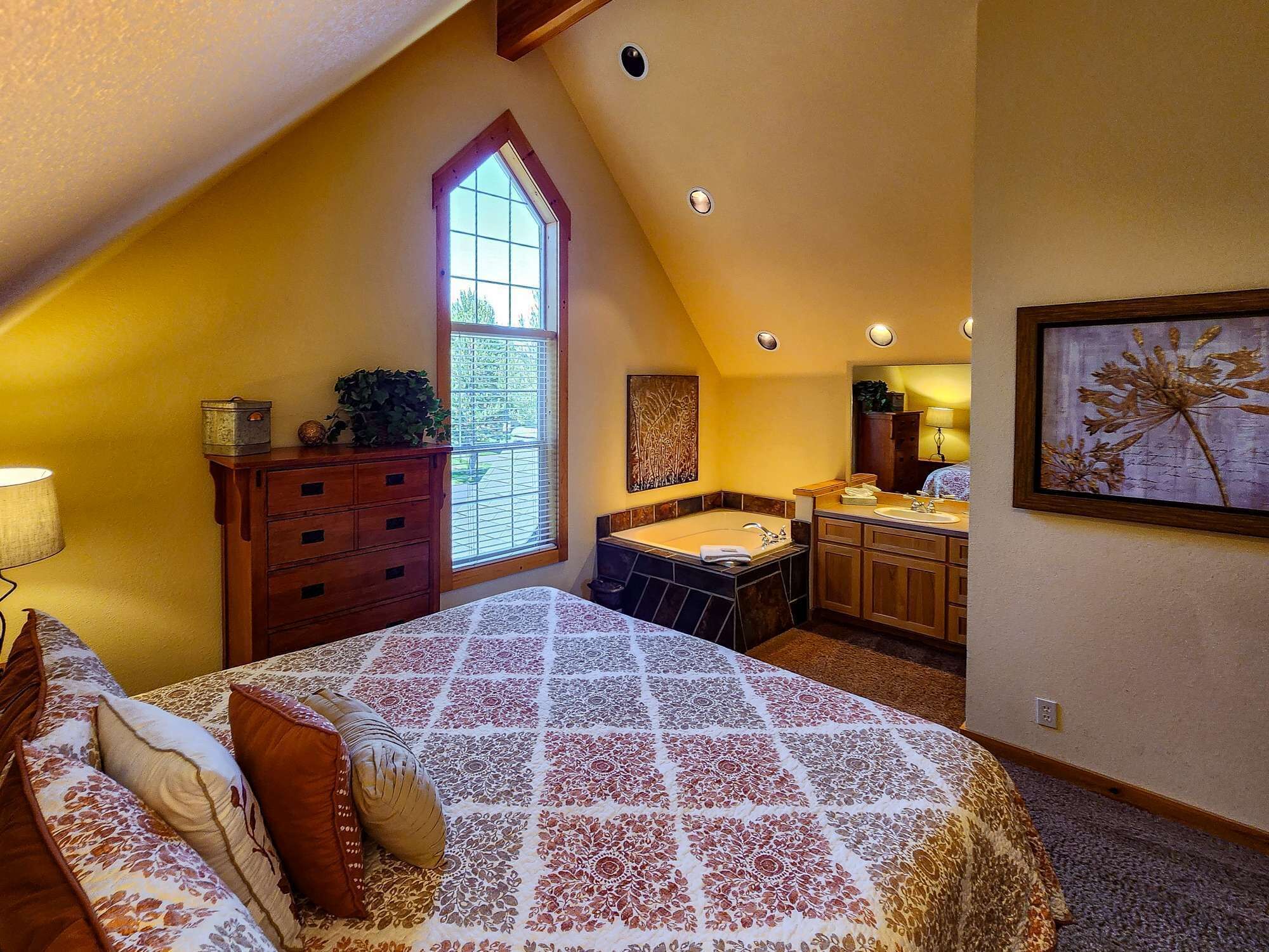 A bedroom with a large bed, a window, a bathroom with a bathtub, and a painting on the wall.