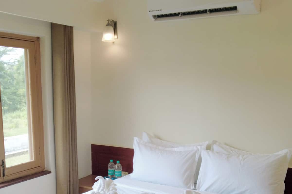 A neatly made bed with white linens, a window with curtains, an air conditioning unit, and a nightstand with two bottles.