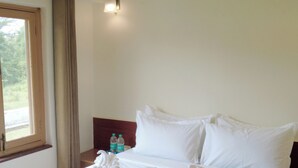 Un lit bien fait, avec du linge de lit blanc, une fenêtre avec des rideaux, un appareil de climatisation et une table de chevet sur laquelle reposent deux bouteilles.