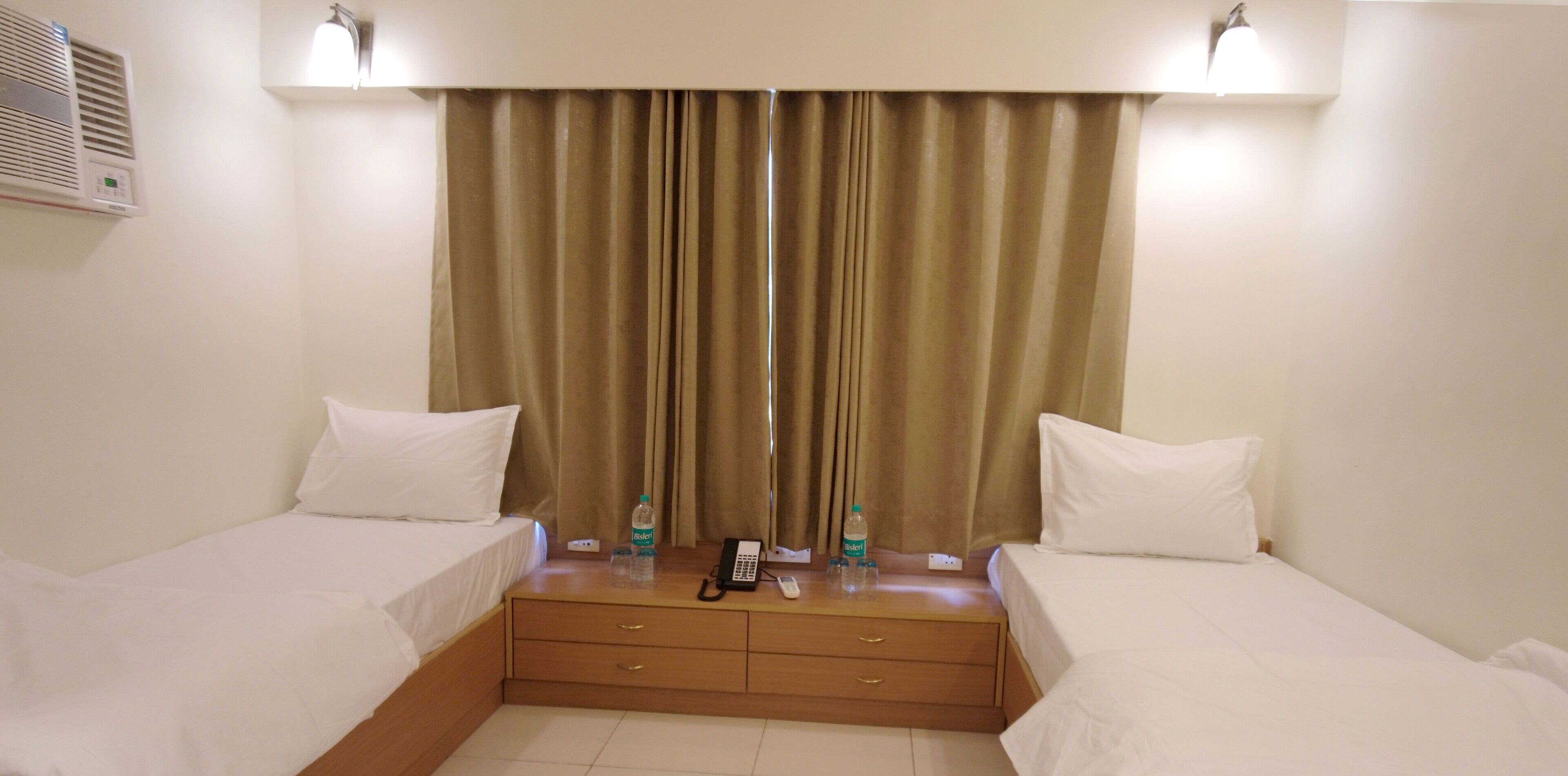 Habitación de hotel con dos camas individuales, una mesita de noche de madera con teléfono y botellas de agua, y un ventanal grande con cortinas.