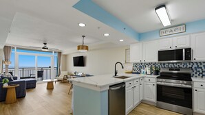 Een moderne keuken met witte kasten, een roestvrijstalen magnetron en een oven. Er is een groot schiereiland met een spoelbak en een vaatwasser. De kamer heeft een blauwe accentmuur en een plafondventilator.