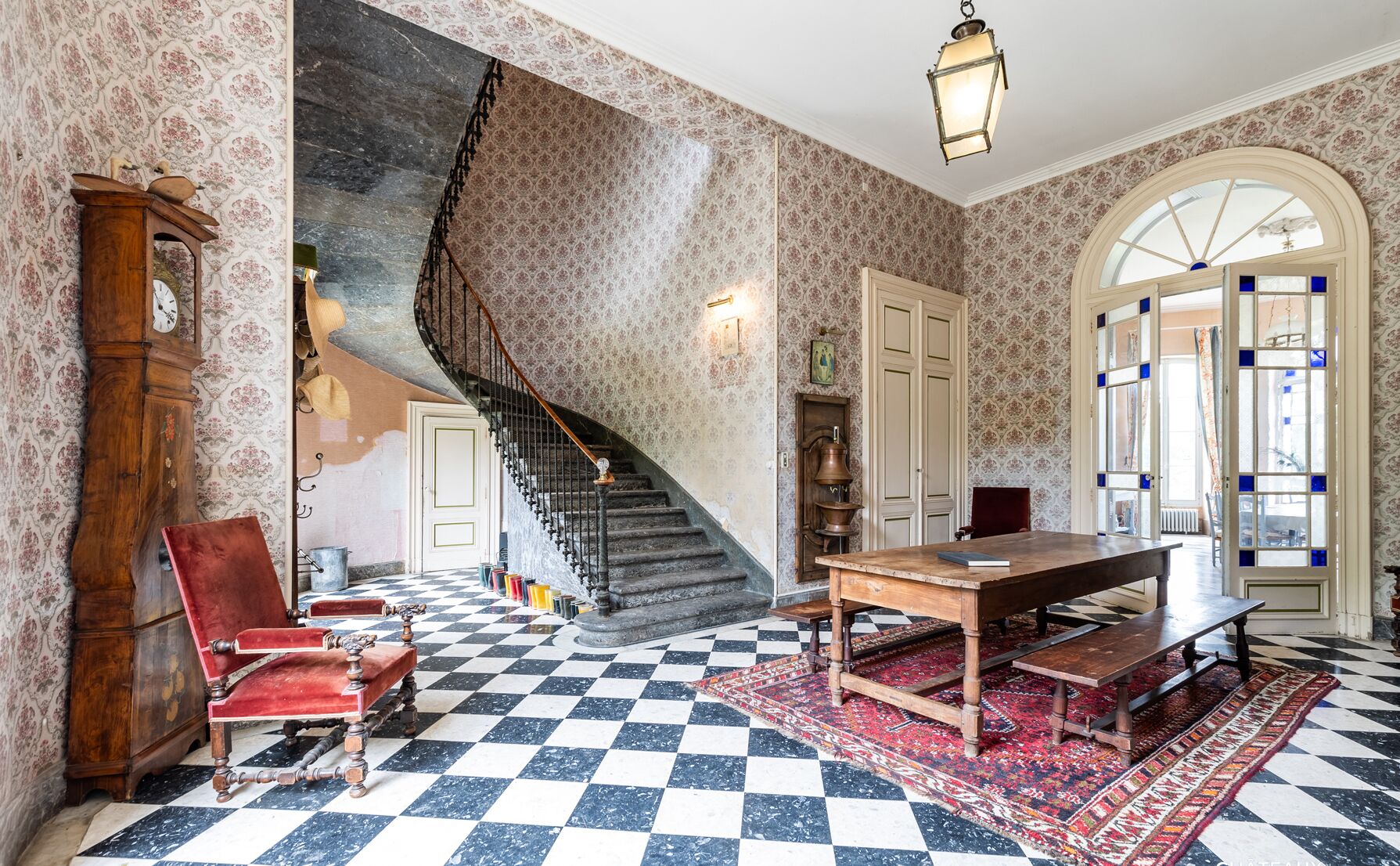 A room with a checkered floor, a wooden table, a red chair, a grandfather clock, and a staircase.