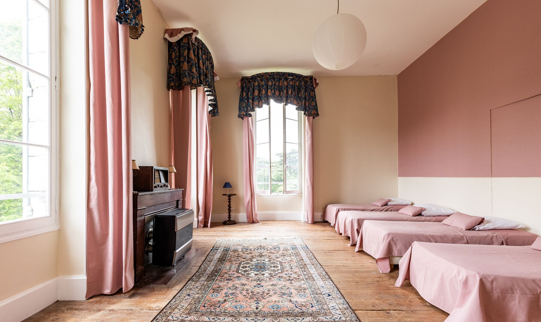 Ein Zimmer mit Holzdielen, einem Teppich in der Mitte, einem Klavier und einer Reihe Betten mit rosa Bettwäsche.