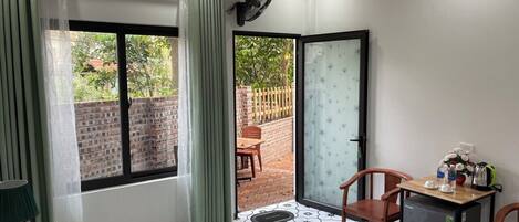 A hotel room with a bed, a desk with a refrigerator, a chair, a table with a vase of flowers, and a view of a brick wall and greenery outside.
