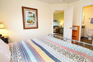 Cozy retreat with a neatly made bed, framed tropical artwork, and a small table with a lamp.