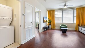 A spacious living room with hardwood flooring, a ceiling fan, and a large window with yellow curtains. - Bright 1BR NOLA Condo with Relaxing Rooftop Pool (New Orleans)