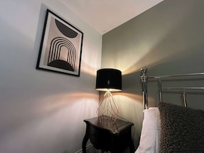 Una habitación con una obra de arte abstracta enmarcada, una lámpara de mesa con base geométrica, un marco de cama de metal y una almohada blanca.
