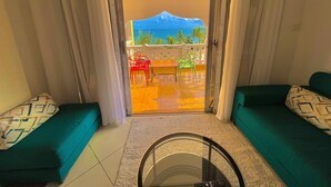 Une pièce dotée d’une porte vitrée donnant sur un balcon avec vue sur la mer et les palmiers. On y trouve deux canapés verts ornés de coussins à motifs, ainsi qu’une petite table avec des chaises.