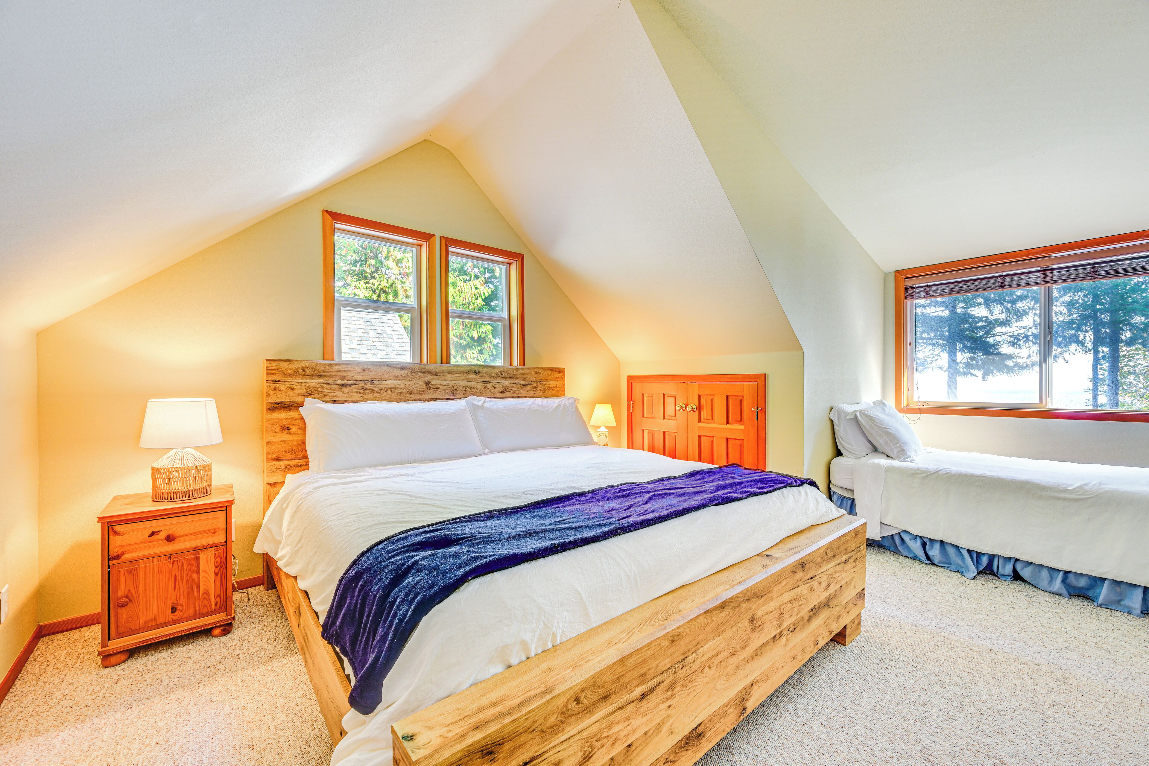 Una camera da letto con soffitto spiovente, un letto grande, un letto più piccolo, comodini in legno e una finestra vista alberi.