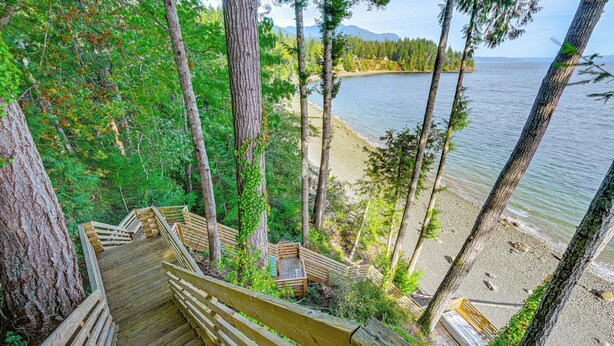 Un escalier en bois descend vers une plage, offrant une vue sur l’eau et la forêt environnante.