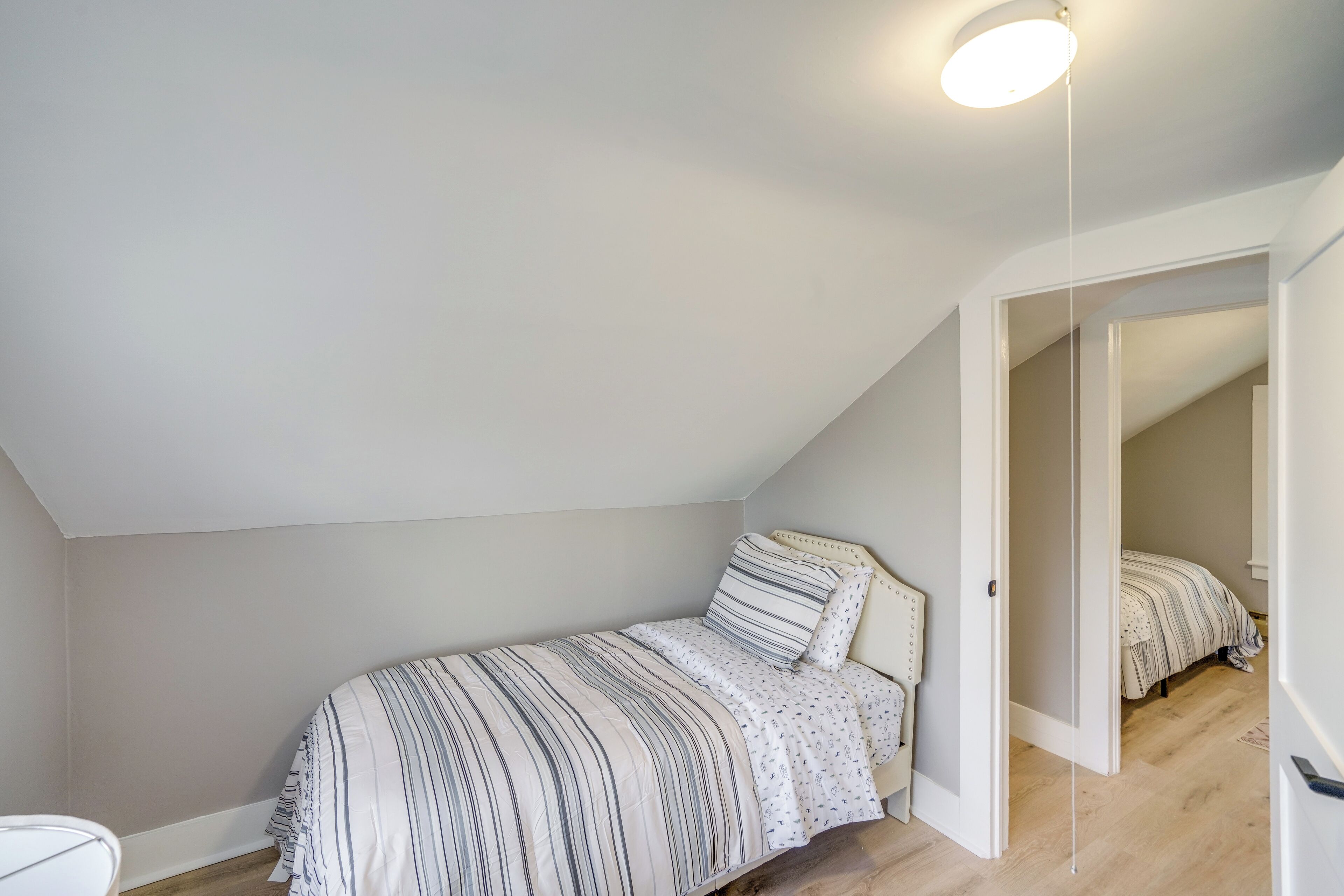 Une chambre avec un plafond en pente, un lit avec une literie à rayures et une porte donnant sur une autre pièce.