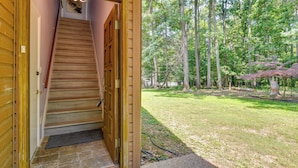 Una escalera de madera conduce a una habitación con piso de piedra, una puerta con felpudo y vista a un patio cubierto de césped y árboles.
