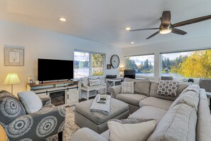 A modern living room with a sectional sofa, a flat-screen TV, a ceiling fan, and large windows offering a view of trees. - 'lakehouse Living' Waterfront Retreat Near Tacoma (Spanaway)
