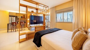 Une chambre d’hôtel moderne dotée d’un grand lit, d’une télévision, d’un bureau avec deux chaises, d’un coin cuisine et offrant une vue sur la ville par la fenêtre.
