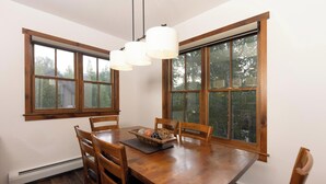 A dining area with a wooden table, four chairs, a chandelier, and large windows with a view of trees. - Aspen Trail Junction: Main St, Walk to Peak 9, Shared Pool & Hot Tub, Shuttle (Breckenridge)