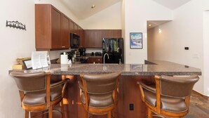 A modern kitchen with dark wood cabinets, a black refrigerator, and a granite countertop. There are three leather bar stools and a framed picture on the wall. - Aspen Trail Junction: Main St, Walk to Peak 9, Shared Pool & Hot Tub, Shuttle (Breckenridge)