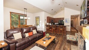 A spacious living room with a leather sofa, wooden coffee table, and a dining area with a wooden table and chairs. A kitchen with dark wooden cabinets and a bar area is visible in the background. - Aspen Trail Junction: Main St, Walk to Peak 9, Shared Pool & Hot Tub, Shuttle (Breckenridge)