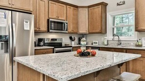 A kitchen with wooden cabinets, stainless steel appliances, granite countertops, and a central island with fruits. - Cascading Falls Retreat by Mountain Oasis | Mountaintown Creek Views (Ellijay)