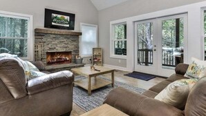 A living room with a stone fireplace, leather couches, and large windows. - Cascading Falls Retreat by Mountain Oasis | Mountaintown Creek Views (Ellijay)