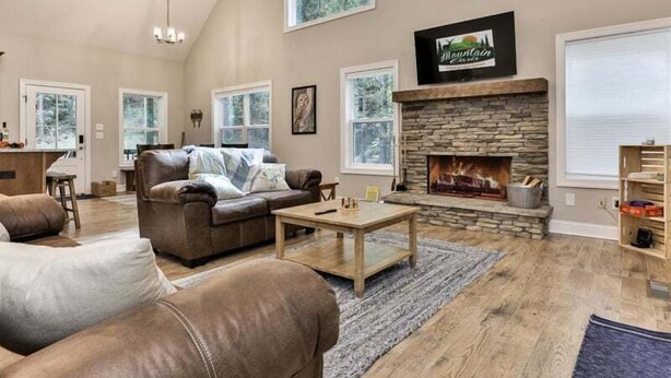 A spacious living room with a stone fireplace, leather sofas, and large windows offering views of the outdoors. - Cascading Falls Retreat by Mountain Oasis | Mountaintown Creek Views (Ellijay)