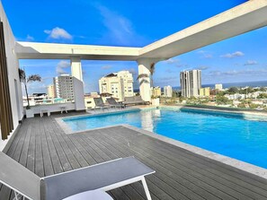 Zona de piscina en la azotea con terraza de madera, sillas de descanso y vistas a la ciudad.