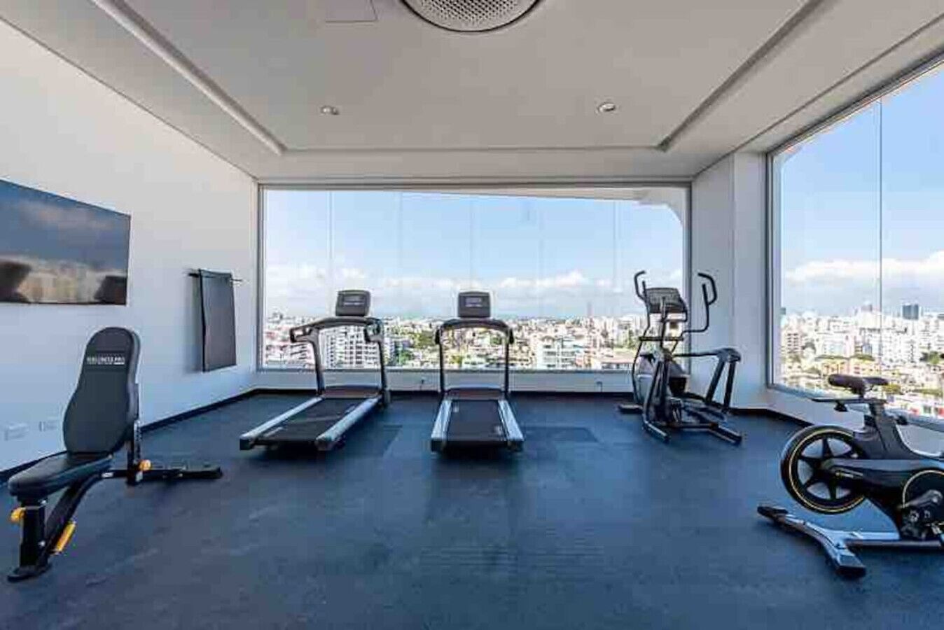 Een sportschool met loopbanden, een crosstrainer en een hometrainer, met stadszicht door grote ramen.