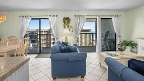 A living room with a sofa, a dining table with chairs, and a view of the outside through large windows. - B515 Charming 2BR Condo | Ocean Views + Steps to Indoor Pool (Myrtle Beach)