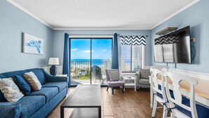 A living room with a sofa, coffee table, and chairs, a TV, and a view of the sea.