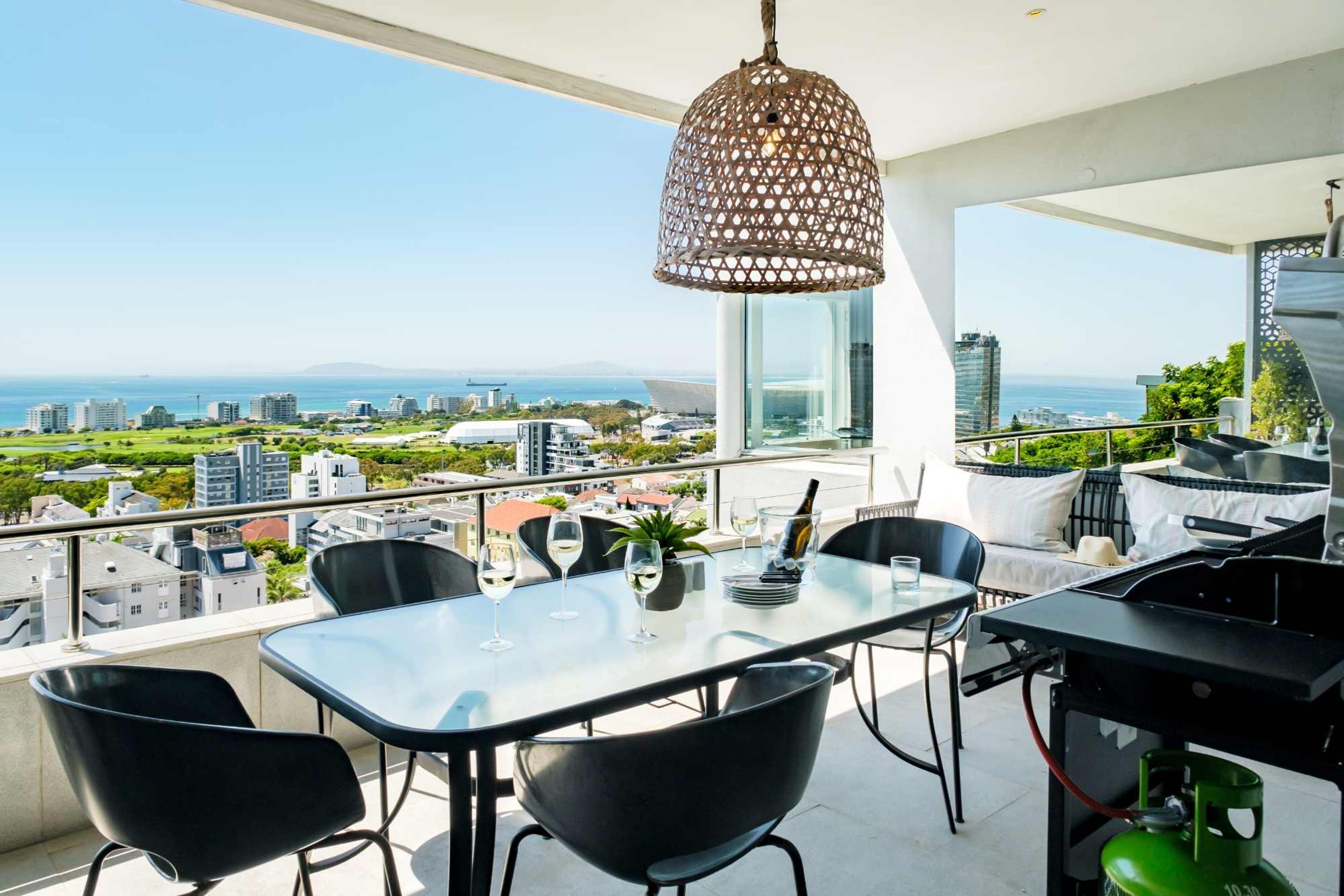 A balcony with a table set for two, a grill, and a view of the city and sea.